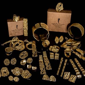 May include: A collection of gold-toned jewellery pieces featuring textured, organic surfaces. The set includes various rings, cuff bracelets, long drop earrings, stud earrings, and necklaces, displayed alongside two brown cardboard boxes with logos.