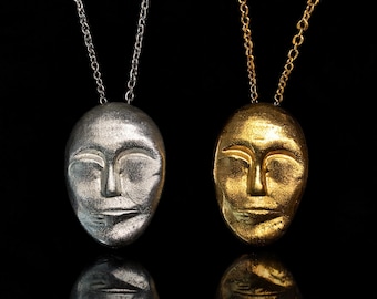 May include: Two necklaces featuring oval-shaped pendants with a sculpted human face design. One pendant is finished in a metallic silver tone on a silver chain, and the other is finished in a metallic gold tone on a matching gold chain.