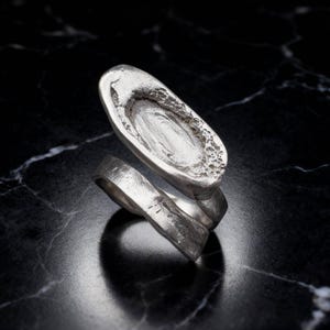 May include: A silver-toned ring featuring an open, wrap-around band design. The top of the ring displays an elongated, oval-shaped focal point with a textured, crater-like surface and a smooth, recessed center, showcasing a raw, metallic finish.