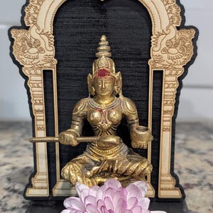 May include: A small golden statue of a Hindu goddess sitting on a throne, with a flower in front of it. The statue is in a wooden frame with intricate carvings.