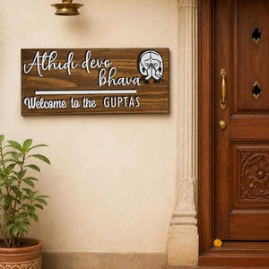 Könnte beinhalten: Ein hölzernes Willkommensschild mit dem Text "Athidi devo bhava" und "Welcome to the GUPTAS" in weißen Buchstaben. Das Schild zeigt eine Grafik einer Person mit gefalteten Händen im Gebet. Das Schild ist an einer beigen Wand angebracht.