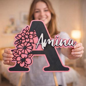 Op de afbeelding: Een houten letter "A" met de naam "Amina" in witte letters. De letter is zwart met een roze omtrek en bloemmotieven. De achtergrond is wazig, wat wijst op een huiselijke omgeving.