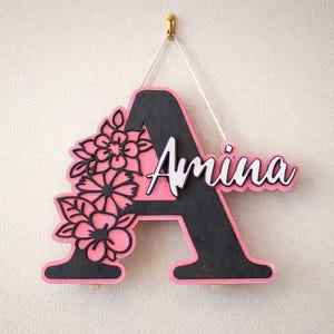 Puede incluir: Adorno de pared de madera rosa y negro con forma de letra "A" con el nombre "Amina" en escritura blanca. La letra está decorada con recortes florales. Se adjunta una cuerda blanca para colgar.