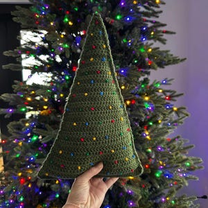 May include: A green crocheted Christmas tree pillow with colorful dots. The pillow is being held in front of a decorated Christmas tree.
