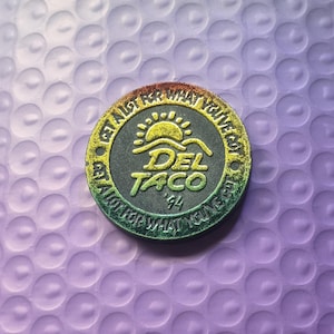 May include: A circular black token with a gradient yellow to green rim. The center features a sun icon above the text DEL TACO '94. The rim text reads GET A LOT FOR WHAT YOU'VE GOT. The design has a textured, metallic finish with raised lettering.