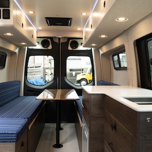 May include: The interior of a converted van with a blue striped bench seat, a table with a wood top, and a kitchen counter with a sink. The van has a white ceiling with LED lights and a white countertop.