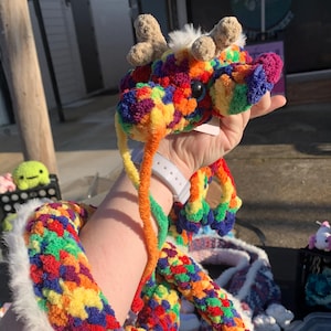 May include: A handmade, colorful crochet dragon with a long, flexible body. The dragon features a rainbow of colors, including red, orange, yellow, green, blue, and purple. It has small, brown horns and white fluffy accents. The dragon is being held up.