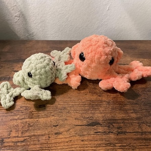 May include: Two plush crochet axolotl toys made from soft yarn. One axolotl is light sage green and the other is a vibrant peach color. Both creatures feature rounded heads, small black plastic eyes, and detailed external gills on the sides.