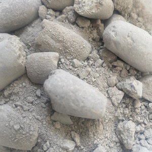 May include: A close-up shot showing a collection of light grey rocks and pebbles. The rocks are of various sizes and shapes, some smooth, some rough. The background is filled with fine, grey particles, creating a textured appearance.