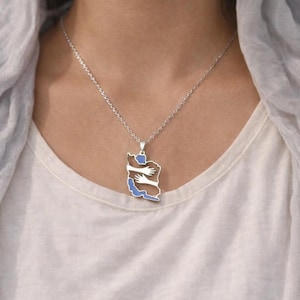 May include: A silver necklace with a pendant shaped like a map of Iran, featuring a blue and brown design with hugging hands. The necklace is worn on a white t-shirt.