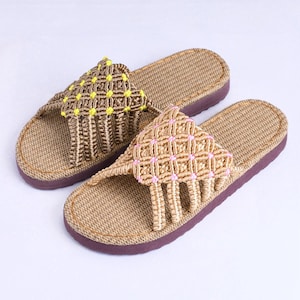 May include: A pair of open-toe slides featuring a woven jute-style footbed and a dark purple sole. The upper strap is crafted from macrame cord in a lattice pattern, with one sandal accented by bright yellow knots and the other by light pink knots.
