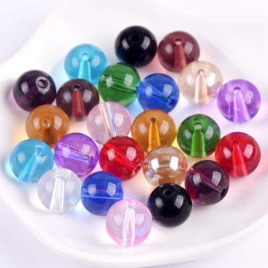 May include: A selection of vibrant, transparent glass beads in shades of blue, green, red, purple, and clear. Each bead features a central hole for threading. The beads are displayed on a white surface.