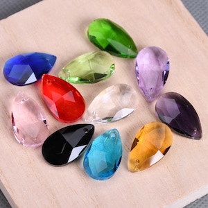 Op de afbeelding: Assortiment van 10 traanvormige kristallen prisma's in verschillende kleuren, waaronder helder, groen, blauw, rood, paars en zwart. De prisma's zijn gefacetteerd en hebben een gat aan de bovenkant om op te hangen.