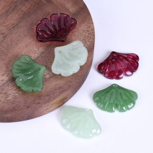 Può includere: Una collezione di ciondoli a forma di ventaglio traslucidi, in pietre preziose, nelle tonalità del verde, verde chiaro e rosso intenso. I ciondoli sono esposti su una superficie di legno, mostrando i loro intricati disegni venati. Ideale per la creazione di gioielli.