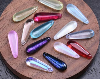 May include: A collection of teardrop-shaped pendants in various finishes, including translucent, opaque, and glitter-infused glass. The assortment features colors such as clear, pink, red, blue, purple, and white, each with a small hole at the top.
