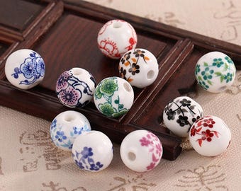 May include: A collection of white ceramic beads with floral designs in various colors, including blue, pink, green, and red. The beads are round with a hole through the center, perfect for jewelry making or crafts. They are displayed on a dark wooden surface.