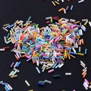 May include: A collection of small, colorful glass beads scattered on a black surface. The beads are cylindrical and come in a variety of colors, including pink, blue, green, yellow, and orange. They are likely used for jewelry making or other crafts.