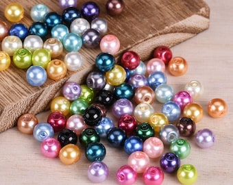 May include: A collection of round, smooth, multicolored faux pearl beads in shades of blue, green, pink, yellow, purple, red, black, white, and brown. Each bead features a central hole for stringing, scattered across a wooden surface.