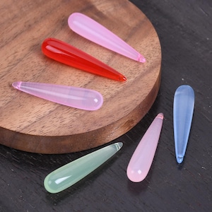 Puede incluir: Una colección de colgantes de vidrio coloridos en forma de lágrima en tonos rojo, rosa, verde y azul. Los colgantes se exhiben sobre una superficie de madera, mostrando su apariencia suave y translúcida. Las formas son alargadas y cónicas.