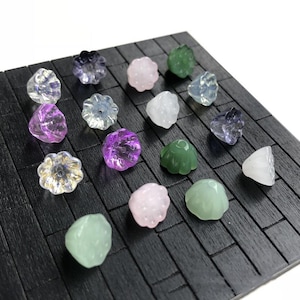 May include: Assortment of 15 small glass flower-shaped buttons in various colours, including pink, green, purple, and clear, displayed on a black surface.