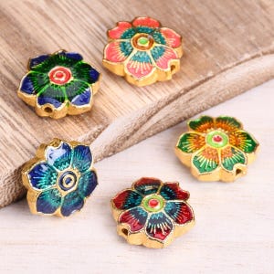 Puede incluir: Cinco cuentas de colores en forma de flor con detalles dorados. Cada cuenta presenta una combinación de colores diferente de rojo, verde, azul y amarillo.