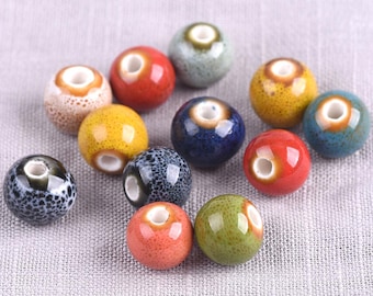 May include: A collection of colorful ceramic beads in various shades, including red, yellow, blue, and green. Each bead has a glossy finish and a white hole. The beads are scattered on a light gray fabric.