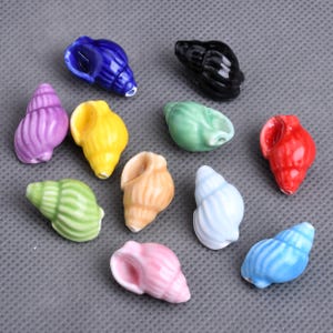 Könnte beinhalten: Eine Sammlung von Keramik-Muscheln in verschiedenen Farben, darunter Blau, Schwarz, Lila, Gelb, Grün, Rot, Hellblau, Rosa und Beige. Die Muscheln sind auf einer grauen Oberfläche angeordnet und zeigen ihre detaillierten Texturen und das glänzende Finish.