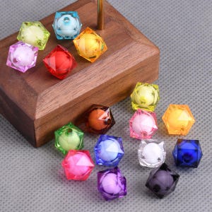 May include: A collection of colorful, faceted beads in various colors including yellow, pink, red, orange, green, blue, and purple. Each bead has a spherical core. The beads are displayed on a wooden surface.