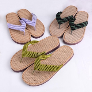 May include: Three pairs of thong sandals featuring tan woven footbeds and dark purple soles. One pair has light purple straps, one pair has black and green striped straps, and one pair has olive green textured straps with a braided appearance.