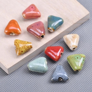 Könnte beinhalten: Eine Sammlung von zehn dreieckigen Keramikperlen in verschiedenen Farben, darunter Orange, Pink, Blau, Gelb, Rot, Weiß, Grün und gesprenkeltes Grau. Jede Perle hat eine glatte, glänzende Oberfläche und ein Loch zum Auffädeln.