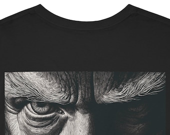 May include: Black t-shirt featuring a detailed, grayscale illustration of a man's face, focusing on the eye and brow. The artwork has a textured, etched appearance, with strong contrast between light and shadow.