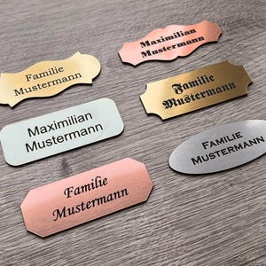 Könnte beinhalten: Sechs verschiedene Metall-Namensschilder mit dem Text "Familie Mustermann" und "Maximilian Mustermann" in schwarzer Schrift. Die Namensschilder sind in verschiedenen Formen und Größen und aus verschiedenen Metallen, darunter Gold, Silber und Kupfer.