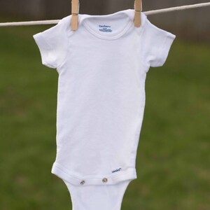 Op de afbeelding: Een witte romper met korte mouwen hangt aan een waslijn met houten wasknijpers. De romper heeft het Gerber-logo en de tekst "onesies" onderaan.