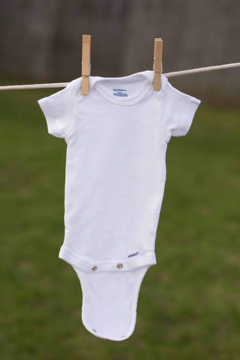 Op de afbeelding: Een witte romper met korte mouwen hangt aan een waslijn met houten wasknijpers. De romper heeft het Gerber-logo en de tekst "onesies" onderaan.
