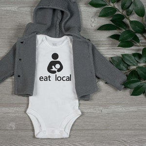 Op de afbeelding: Witte baby romper met een zwarte afbeelding van een zogende moeder en de tekst "eat local". De romper ligt op een grijs houten oppervlak met een groene, bladerrijke tak.