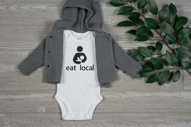 Op de afbeelding: Witte baby romper met een zwarte afbeelding van een zogende moeder en de tekst "eat local". De romper ligt op een grijs houten oppervlak met een groene, bladerrijke tak.