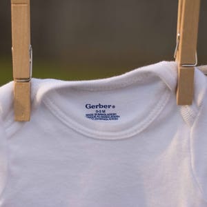 Op de afbeelding: Een witte baby romper hangend aan een waslijn met houten wasknijpers. De romper heeft een blauw Gerber label met de tekst "Gerber&reg; 0-3 M MADE IN BANGLADESH 100% COTTON" erop gedrukt.