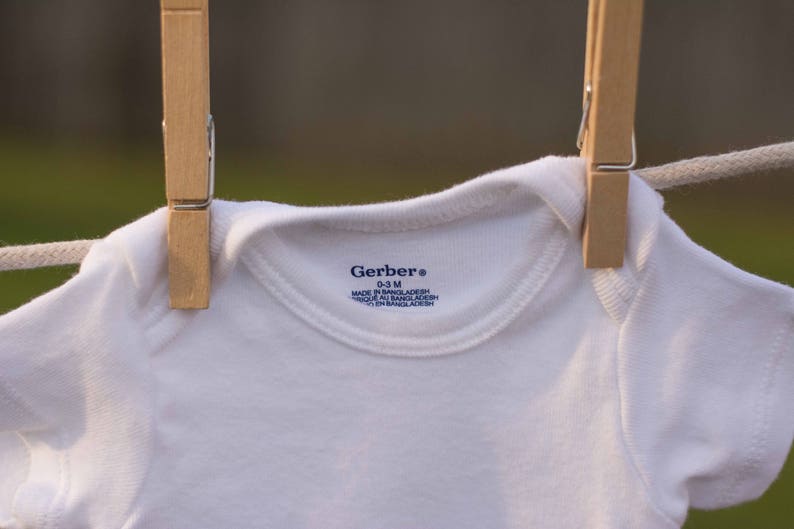 Op de afbeelding: Een witte baby romper hangend aan een waslijn met houten wasknijpers. De romper heeft een blauw Gerber label met de tekst "Gerber&reg; 0-3 M MADE IN BANGLADESH 100% COTTON" erop gedrukt.