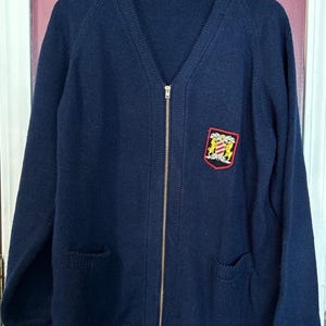 May include: Navy blue zip-up cardigan sweater with a V-neck and two front pockets. The sweater features a gold zipper and a crest patch on the left chest. The crest is red, gold, and black.