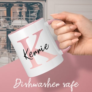 May include: White ceramic mug with a pink interior and handle. The mug features a large pink letter "K" and the name "Kerrie" in black script. The words "Dishwasher safe" are at the bottom of the image.