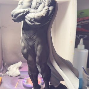 May include: A gray and white statue of a muscular figure with a white cape. The statue has a detailed face with a mustache and is standing on a textured base. The figure's arms are crossed.