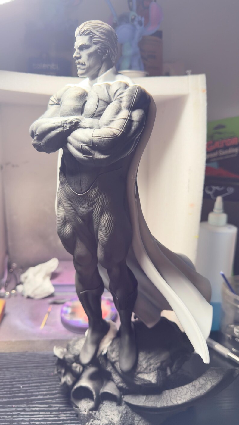 May include: A gray and white statue of a muscular figure with a white cape. The statue has a detailed face with a mustache and is standing on a textured base. The figure's arms are crossed.