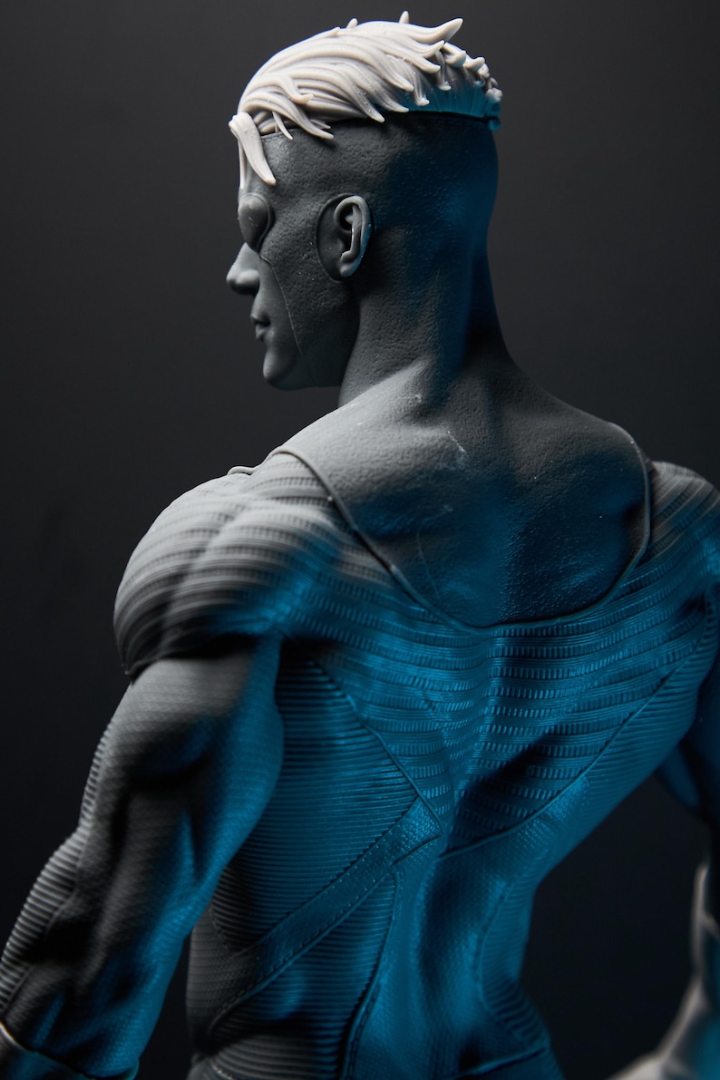 May include: A gray figurine of a superhero, viewed from the back and side. The figure has a muscular physique, with detailed textures on the suit and hair. The lighting creates shadows, highlighting the form and design.