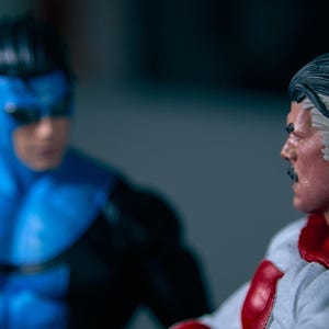 May include: Two action figures of comic book characters. One figure is in a blue and black suit with a mask. The other figure is in a white and red suit with a cape and a mustache. The figures are facing each other.