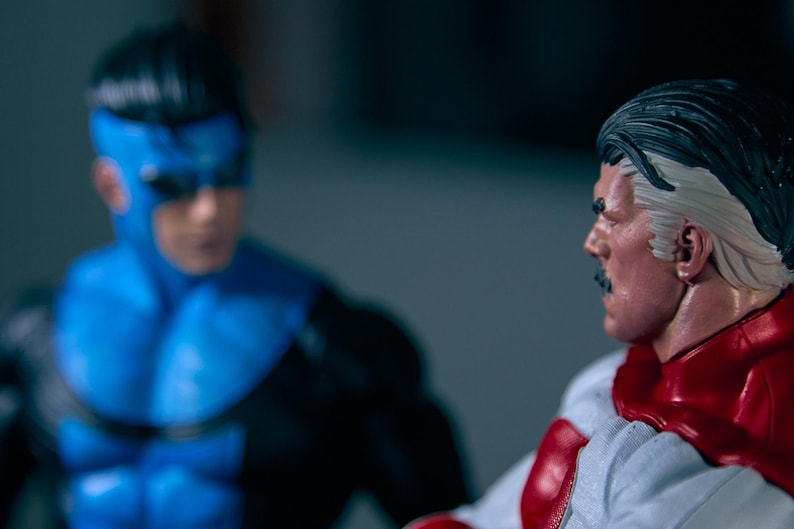 May include: Two action figures of comic book characters. One figure is in a blue and black suit with a mask. The other figure is in a white and red suit with a cape and a mustache. The figures are facing each other.