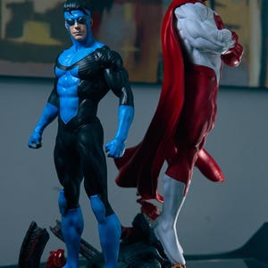 May include: Two superhero action figures. One figure is in a blue and black suit with a mask. The other figure is in a red and white suit with a red cape. Both figures are standing on a rocky base.