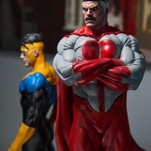 May include: Two superhero action figures. The primary figure is in a red and gray suit with a red cape, arms crossed. The second figure is in a yellow, blue, and black suit. The figures are highly detailed and muscular.