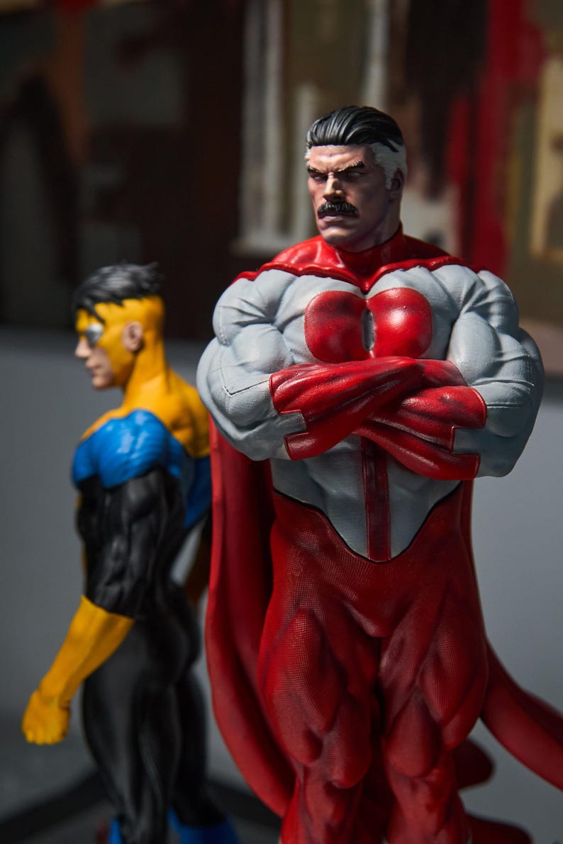 May include: Two superhero action figures. The primary figure is in a red and gray suit with a red cape, arms crossed. The second figure is in a yellow, blue, and black suit. The figures are highly detailed and muscular.