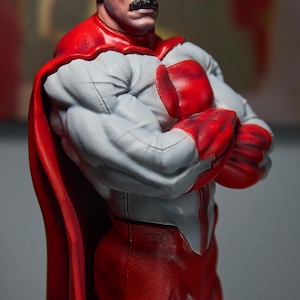 May include: A detailed figurine of a muscular superhero with a stern expression. The figure wears a gray and red costume with a red cape. The character has a distinctive mustache and white hair streaks.
