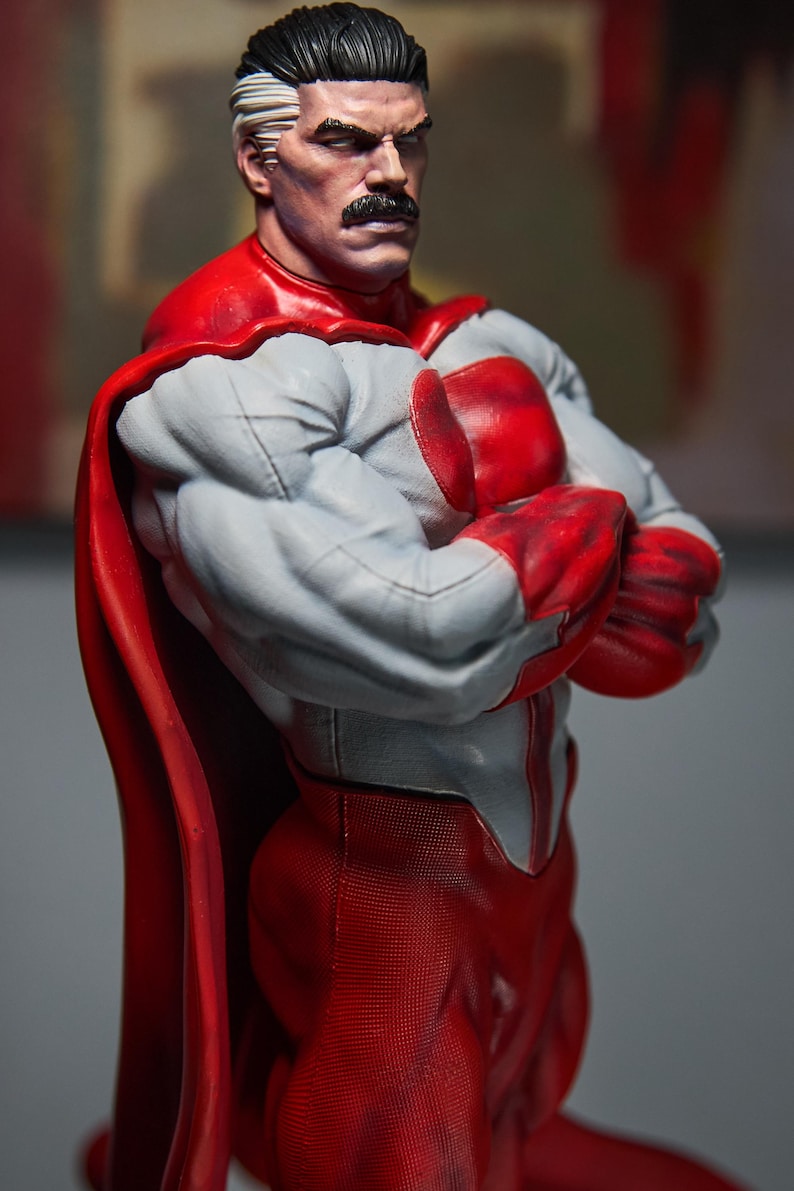 May include: A detailed figurine of a muscular superhero with a stern expression. The figure wears a gray and red costume with a red cape. The character has a distinctive mustache and white hair streaks.